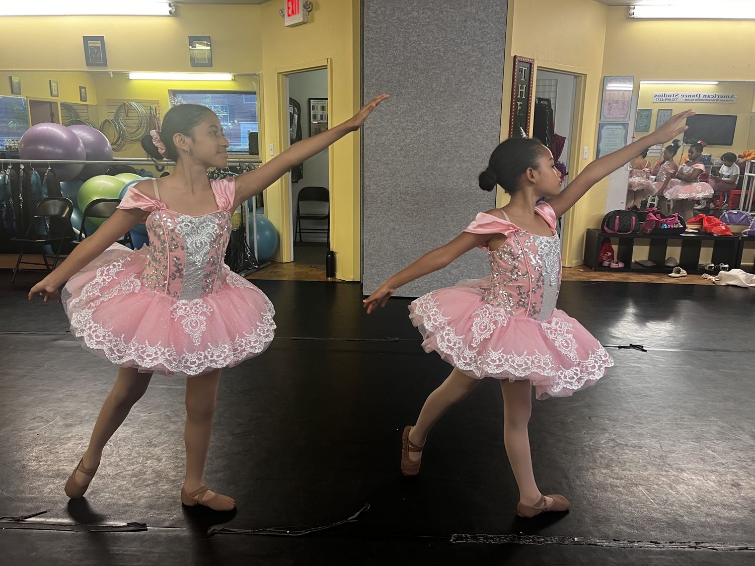Two ballet students in pink lace tutus posing in arabesque arms in the studio