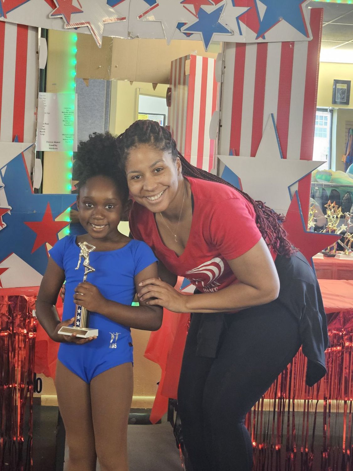 A young dancer in a blue leotard holding her graduation trophy with her teacher under the star arch