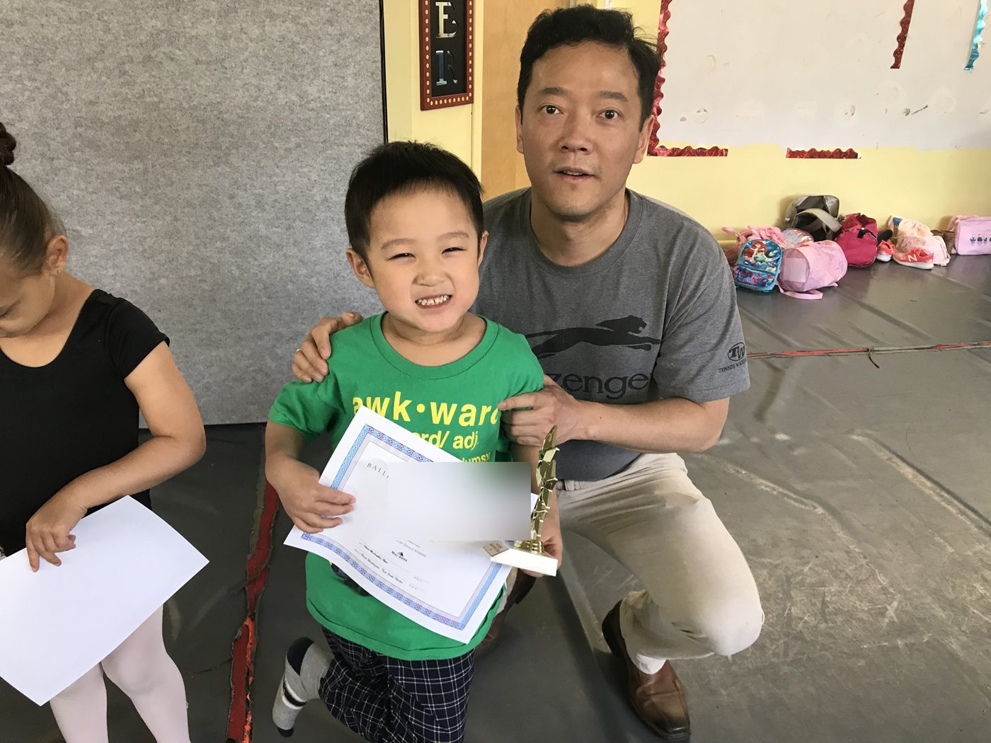 A proud dad kneeling beside his son, who holds his syllabus certificate and trophy