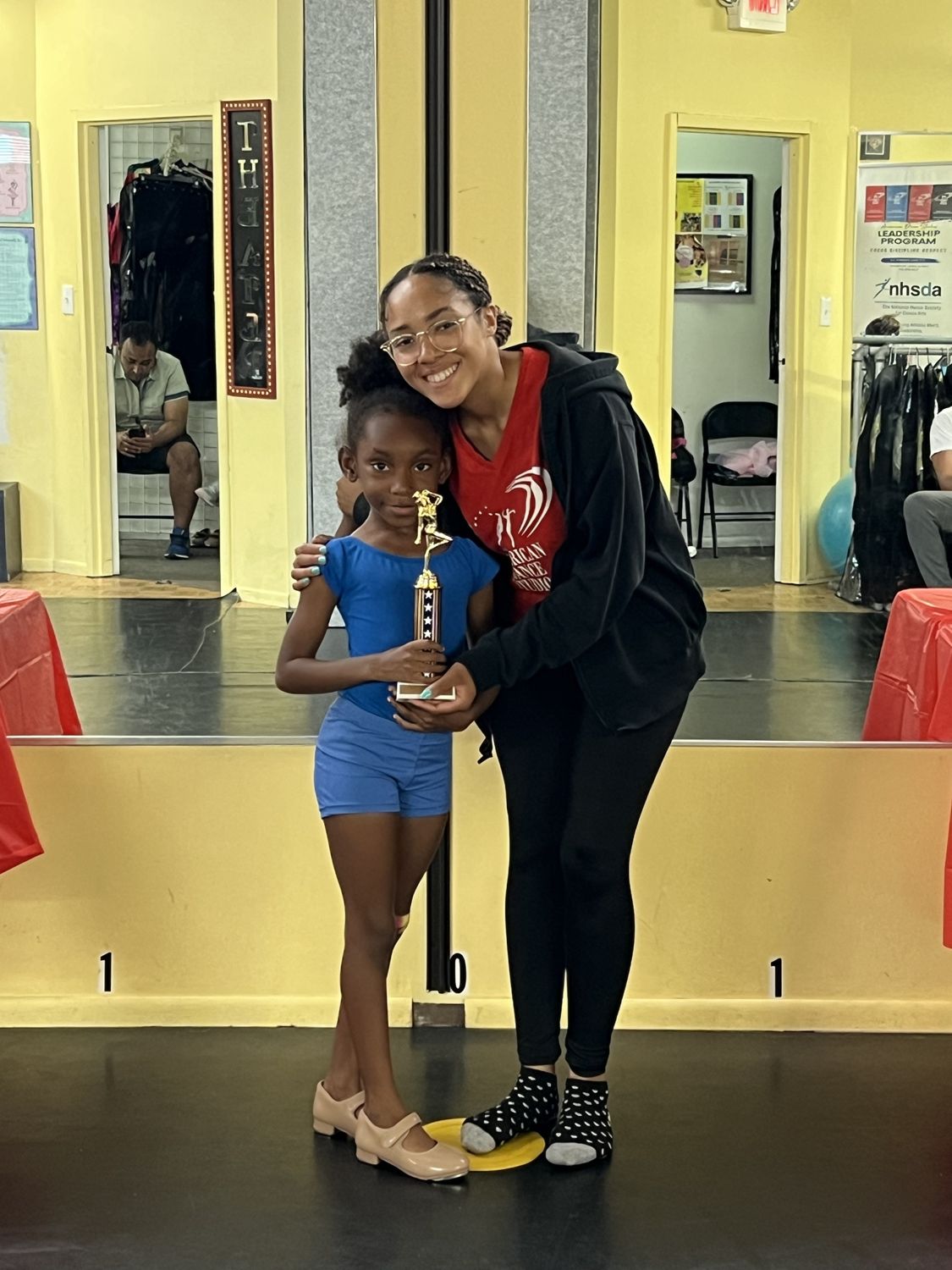 A teacher hugging a young dancer holding her trophy