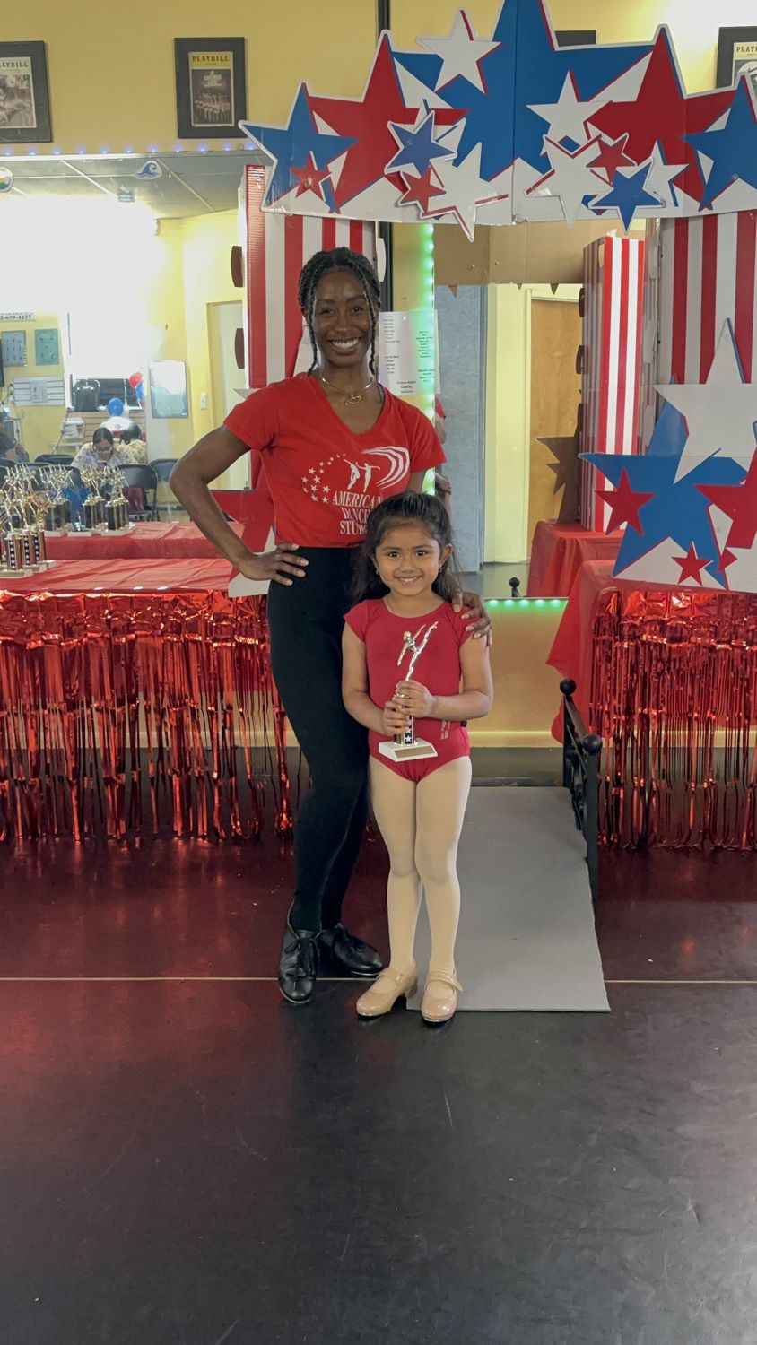 A young tap dancer holding her graduation trophy with her teacher in front of the star arch