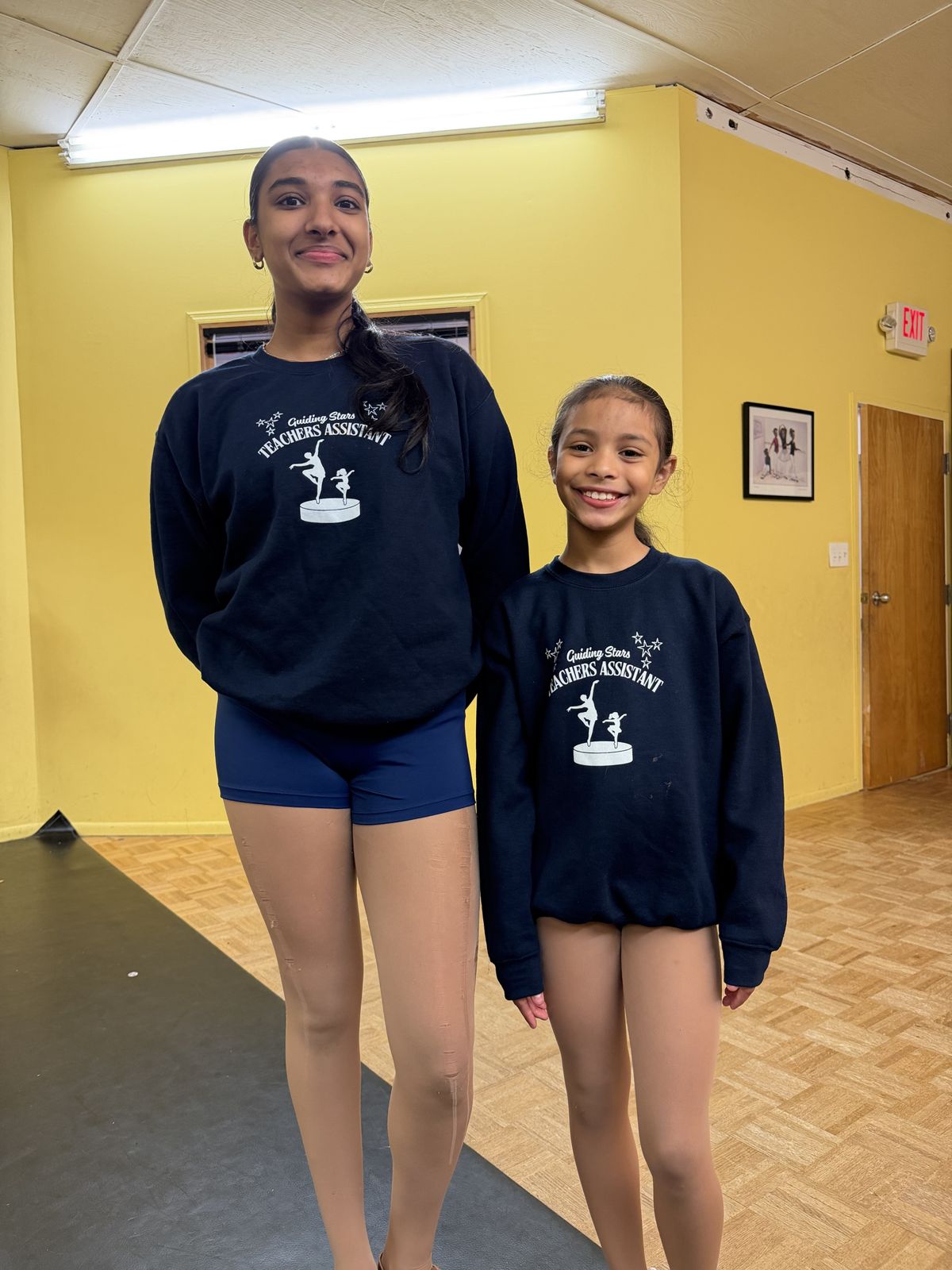 An older and a younger teacher assistant side by side in matching sweatshirts