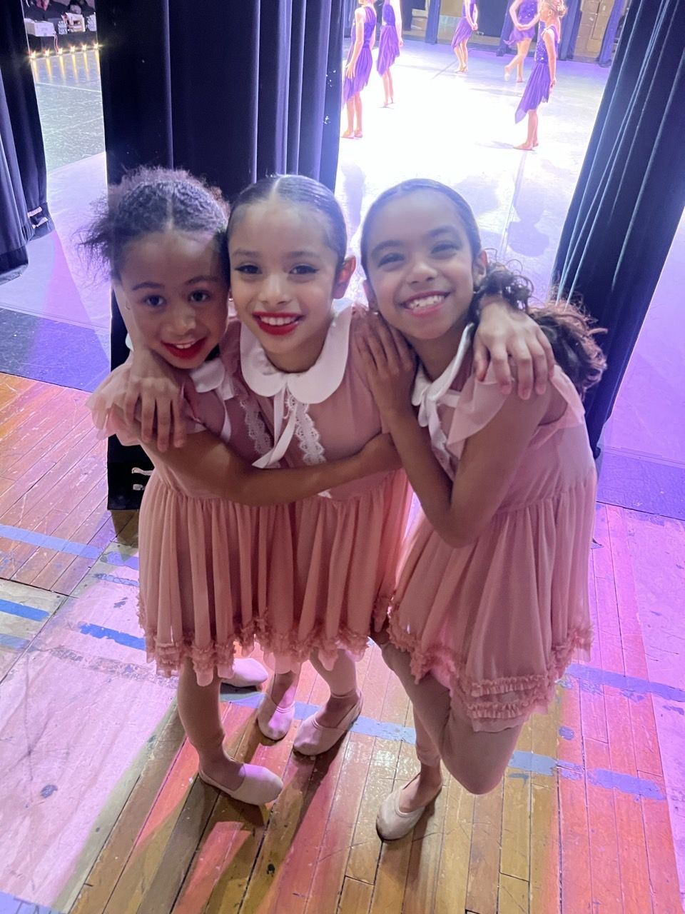 Three young dancers in pink dresses hugging backstage