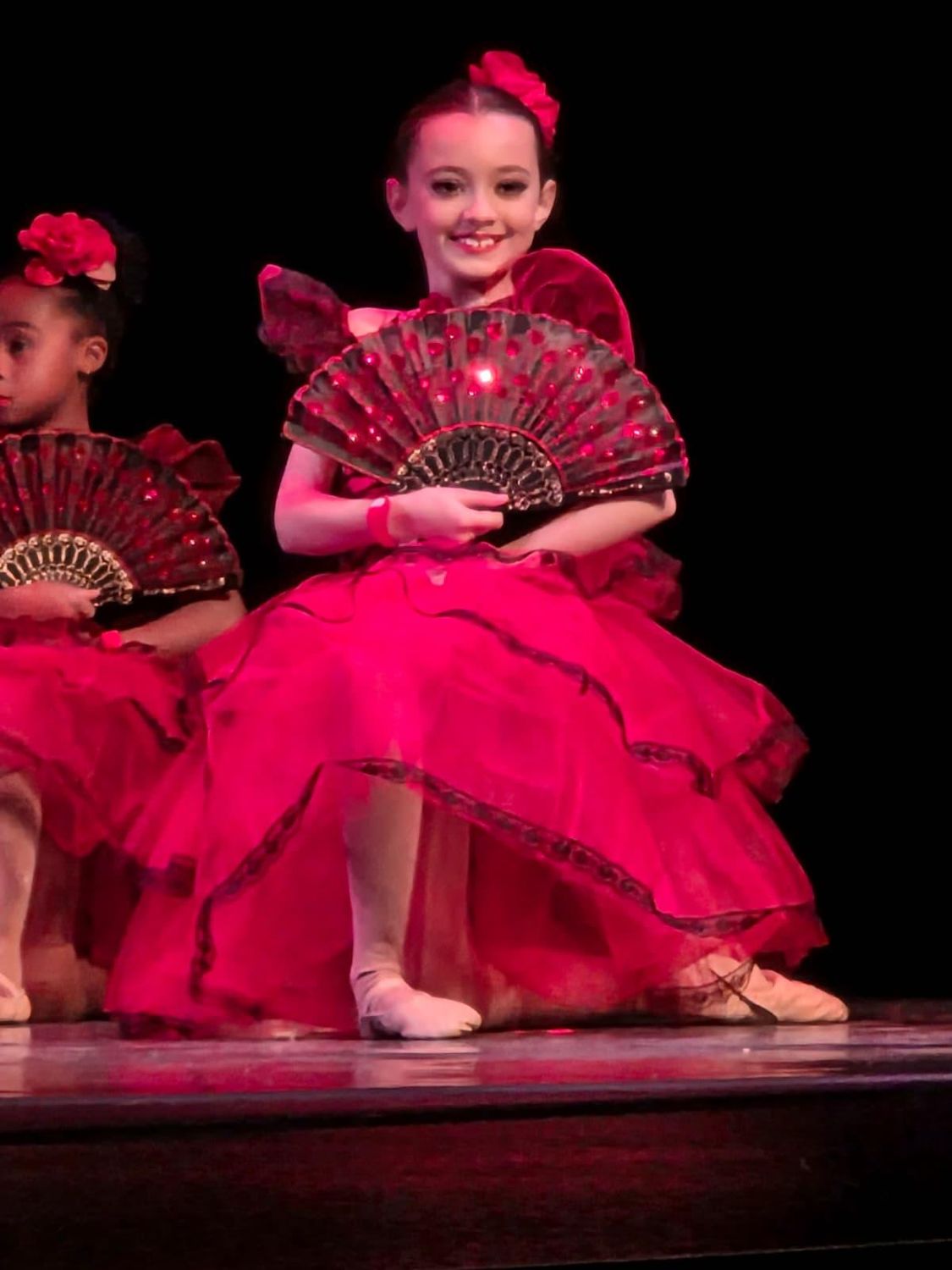 A young dancer smiling on stage with a red Spanish fan